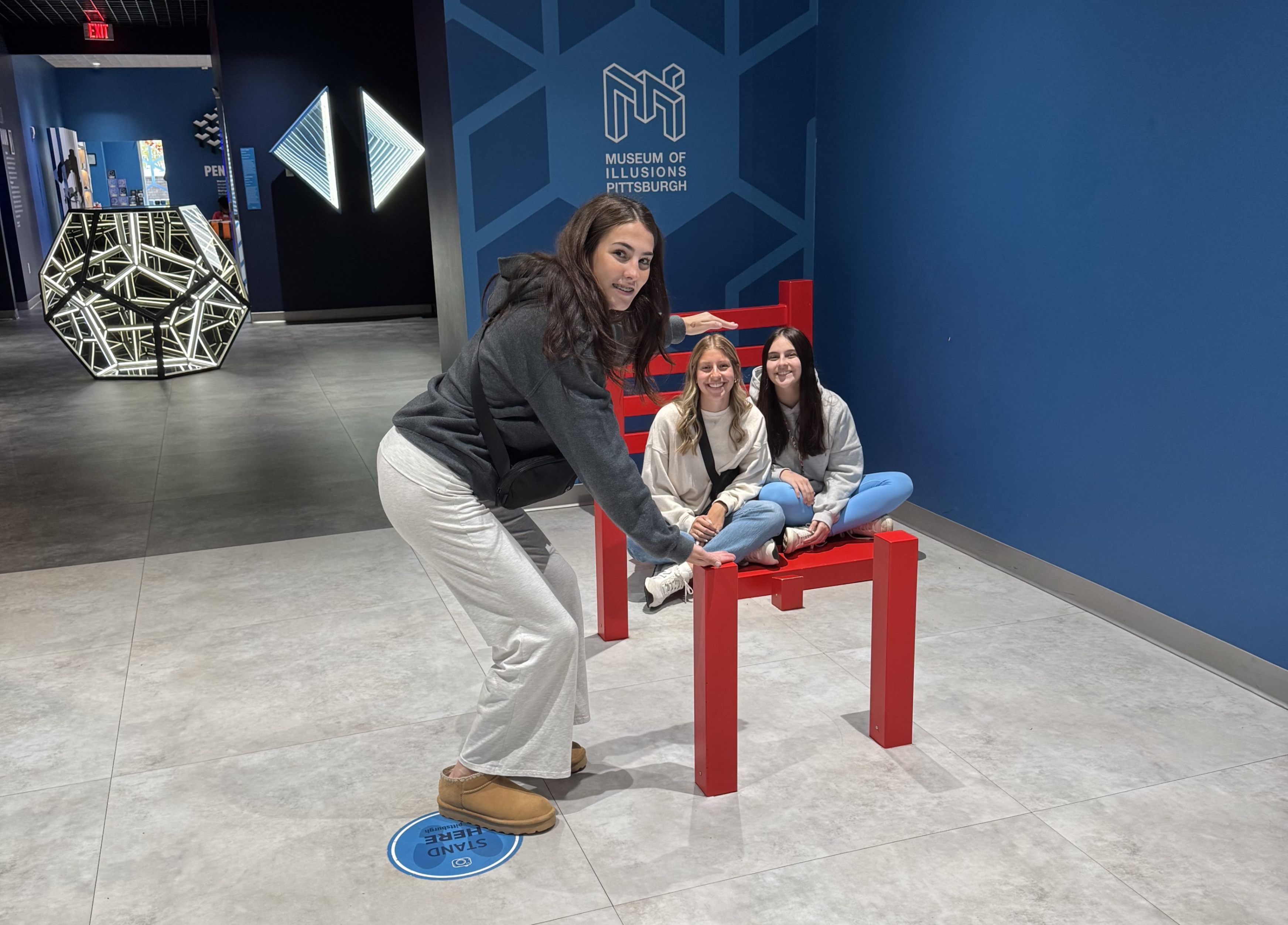 Students pose for a photo at the museum of illusions that makes one student look much larger than the other two.